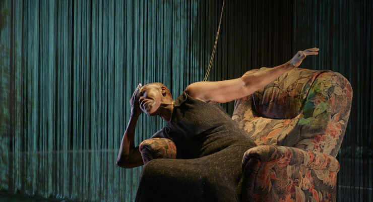 Frau in langem Kleid lehnt mit ausgestrecktem Arm auf einem gemusterten Sessel vor einem Hintergrund aus hängenden Fäden