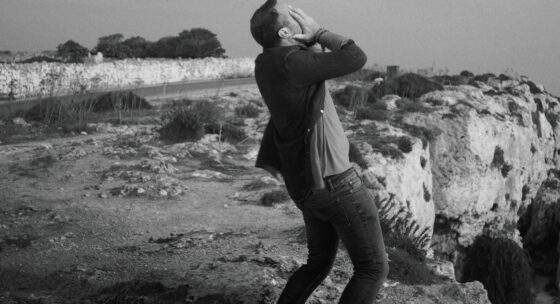 Ein Mann steht auf felsigem Gelände an einer Küste und hält sich mit beiden Händen das Gesicht, während er sich nach hinten lehnt.