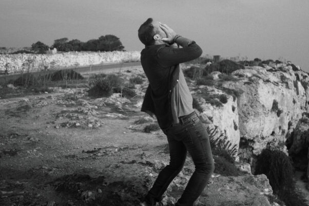 Ein Mann steht auf felsigem Gelände an einer Küste und hält sich mit beiden Händen das Gesicht, während er sich nach hinten lehnt.