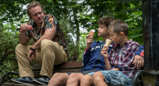 Drei Personen sitzen auf einer Parkbank, zwei Kinder essen Eis, ein Erwachsener sitzt mit angewinkeltem Arm daneben.