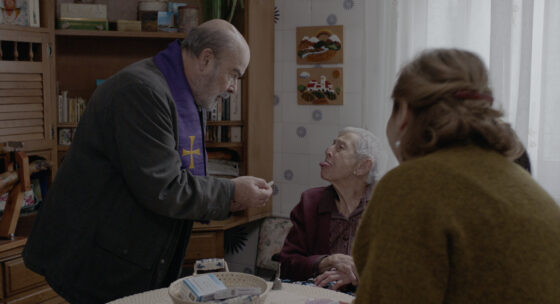 Drei Personen sitzen und stehen an einem Küchentisch in einem Raum mit gemusterter Tapete und Regalen im Hintergrund