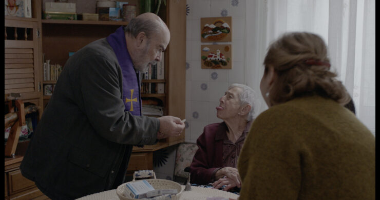 Drei Personen sitzen und stehen an einem Küchentisch in einem Raum mit gemusterter Tapete und Regalen im Hintergrund