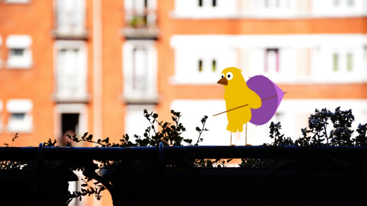 Gelbe Comicfigur mit großem Auge und lila Regenschirm steht auf einem Zaun vor einem unscharfen Wohnhaus.