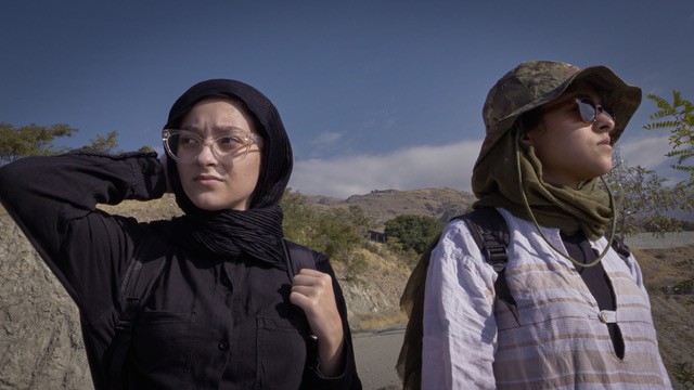 Zwei Personen mit Kopfbedeckungen und Schals stehen im Freien vor einer bergigen Landschaft unter blauem Himmel.