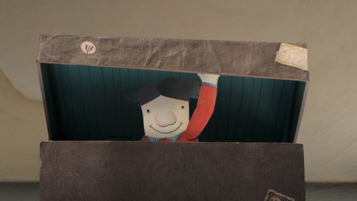 Kind mit rotem Pullover hebt den Arm und schaut aus einer geöffneten, braunen Kiste heraus.