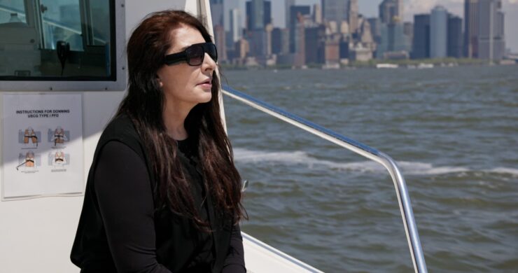 Frau mit langen dunklen Haaren und schwarzem Oberteil sitzt auf einem Boot mit Wasser und Stadt im Hintergrund