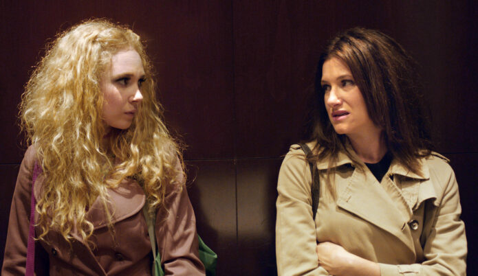 Zwei Frauen mit langen Haaren sitzen nebeneinander auf einer dunklen Ledercouch, eine trägt einen hellen Trenchcoat, die andere einen braunen Pullover.