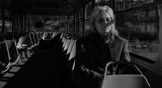 Ältere Person mit hellem Haar sitzt allein in einem fast leeren Bus mit Reihen von Sitzplätzen auf beiden Seiten