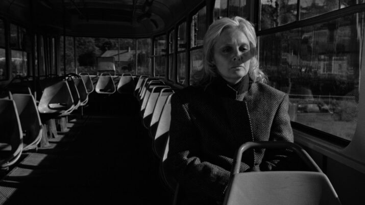 Ältere Person mit hellem Haar sitzt allein in einem fast leeren Bus mit Reihen von Sitzplätzen auf beiden Seiten