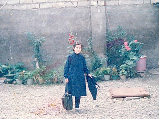 Person in dunklem Mantel steht auf einem kiesbedeckten Hof vor einer Mauer mit mehreren Blumentöpfen und blühenden Pflanzen