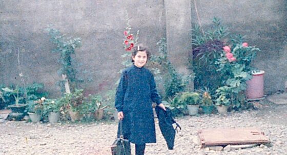 Person in dunklem Mantel steht auf einem kiesbedeckten Hof vor einer Mauer mit mehreren Blumentöpfen und blühenden Pflanzen