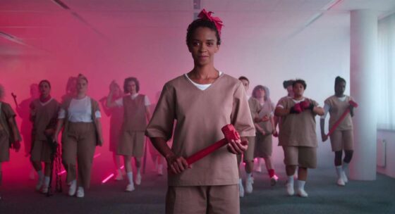 Gruppe von Personen in beigen Overalls in einem Raum mit rosa Licht, vorne eine Person mit rotem Hammer in den Händen