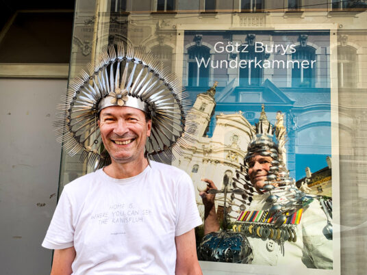 Eine Person mit auffälliger Kopfbedeckungen aus Metall vor einem Schaufenster mit der Aufschrift 'Götz Burys Wunderkammer' und einem Bild.
