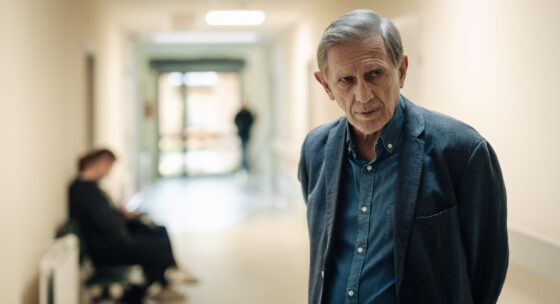 Älterer Mann mit grauen Haaren trägt dunkelblauen Blazer und hellblaues Hemd in einem hellen Flur mit Sitzbank und einer weiteren Person im Hintergrund.