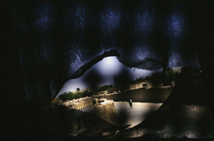 Blick durch einen zerrissenen Schleier auf eine von Lehmwänden umgebene Straße mit einer einzelnen Person im Hintergrund