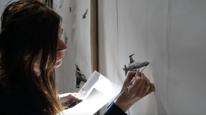 Frau mit dunklen Haaren hält Pinsel und Papier und malt an einer weißen Wand mit kleinen schwarzen Flugzeugzeichnungen