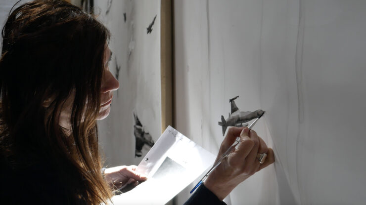 Frau mit dunklen Haaren hält Pinsel und Papier und malt an einer weißen Wand mit kleinen schwarzen Flugzeugzeichnungen