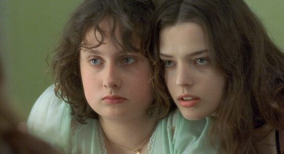 Zwei Frauen mit langen dunklen Haaren sitzen nebeneinander, eine trägt eine weiße Bluse und eine goldene Halskette