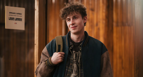 Junge mit lockigem Haar trägt eine Jacke mit Ärmeln in zwei Farben und hält einen Rucksackriemen über der Schulter