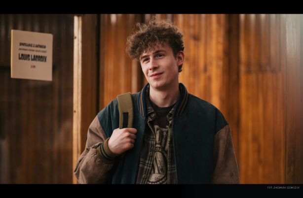 Junge mit lockigem Haar trägt eine Jacke mit Ärmeln in zwei Farben und hält einen Rucksackriemen über der Schulter