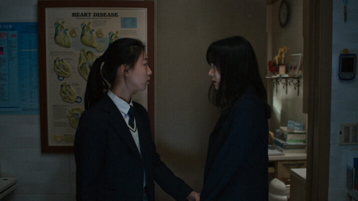 Zwei dunkel gekleidete Personen stehen sich in einem schwach beleuchteten Raum gegenüber, im Hintergrund ein Poster mit Herzdiagrammen.