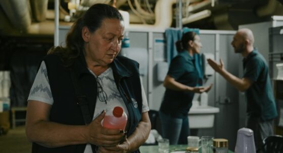 Frau hält Flasche in einem Raum mit Tisch und Geschirr, im Hintergrund streiten sich zwei Personen.