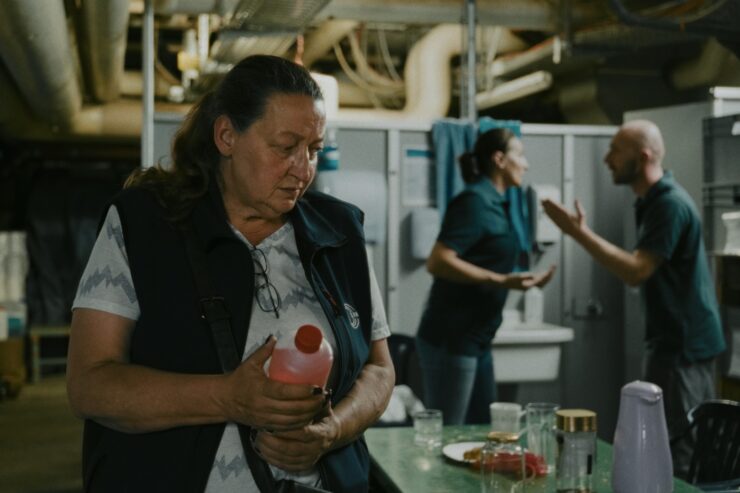 Frau hält Flasche in einem Raum mit Tisch und Geschirr, im Hintergrund streiten sich zwei Personen.