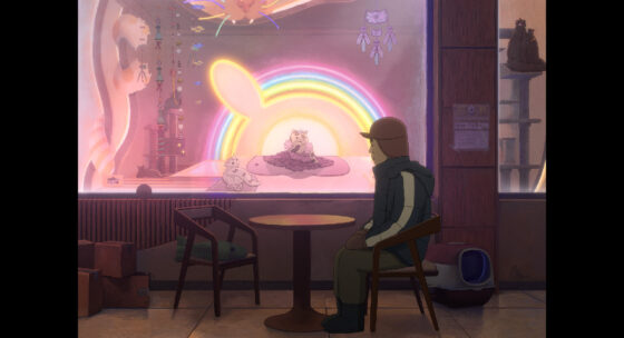 Person mit Brille und Mütze sitzt an einem runden Tisch vor einem Schaufenster mit einer leuchtenden Regenbogen-Dekoration und ausgestellten Stofftieren