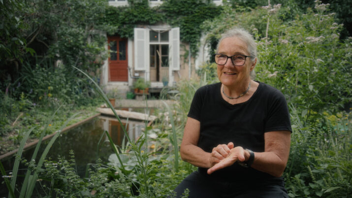 Ältere Person mit kurz geschnittenem grauem Haar und schwarzem T-Shirt sitzt in einem Garten vor einem Teich und einem Haus mit weißen Fensterläden