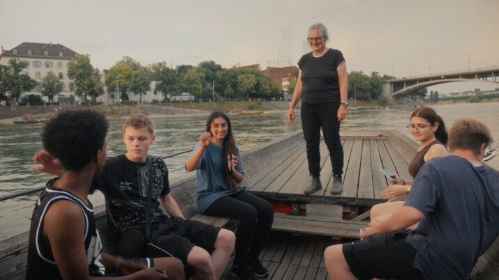 Gruppe von sechs Personen sitzt und steht auf einem Holzsteg am Flussufer mit Brücke und Gebäuden im Hintergrund