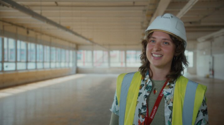 Person mit Bauhelm, Warnweste und rotem Ausweisband in einem großen, leeren Raum mit vielen Fenstern