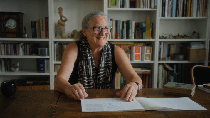 Ältere Frau mit grauen Haaren und schwarzem, gemustertem Schal sitzt an einem Holztisch vor einem aufgeschlagenen Buch in einem Raum mit Bücherregalen im Hintergrund