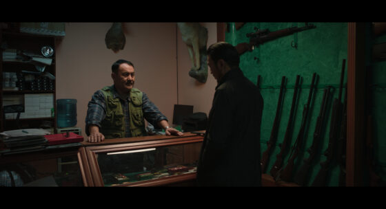 Zwei Männer stehen sich in einem Raum mit Waffen an der Wand gegenüber, einer lehnt sich an einen Glastresen mit ausgestellten Messern.