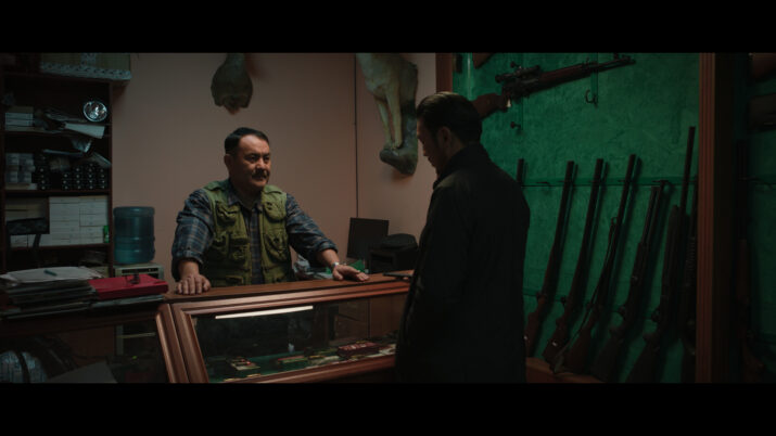 Zwei Männer stehen sich in einem Raum mit Waffen an der Wand gegenüber, einer lehnt sich an einen Glastresen mit ausgestellten Messern.