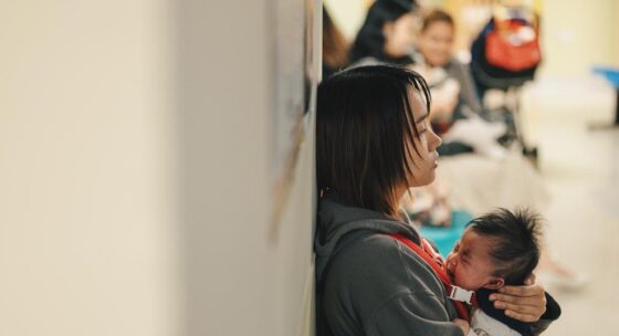 Frau sitzt mit Baby auf dem Arm an einer Wand in einem öffentlichen Raum