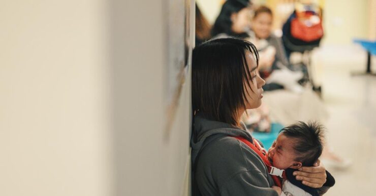 Frau sitzt mit Baby auf dem Arm an einer Wand in einem öffentlichen Raum