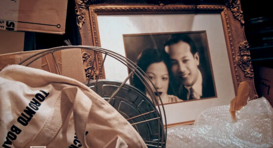 Ein vergoldeter Bilderrahmen mit einem unscharfen Porträtfoto steht in einem Keller zwischen Kartons und einer Stofftasche mit der Aufschrift 'TORONTO BOARD'.