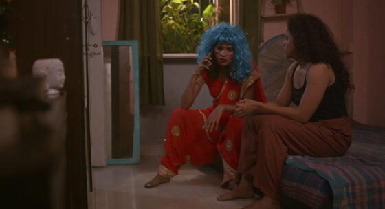 Zwei Frauen sitzen in einem Zimmer, eine mit blauer Perücke und rotem Kleid auf dem Boden, die andere mit dunklen Haaren und brauner Hose auf einem Bett