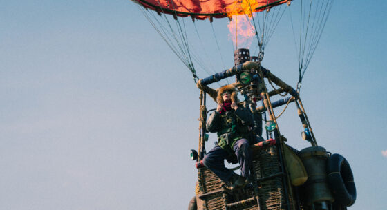 Person in warmer Kleidung sitzt in einem Heißluftballonkorb mit brennender Flamme über dem Korb