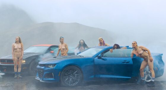 Sechs nackte Frauen umgeben zwei Sportwagen, einen blauen und einen schwarzen, in nebliger Berglandschaft