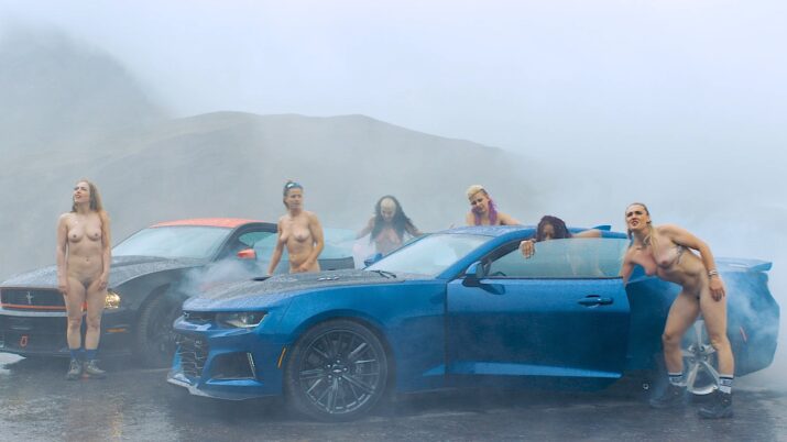 Sechs nackte Frauen umgeben zwei Sportwagen, einen blauen und einen schwarzen, in nebliger Berglandschaft
