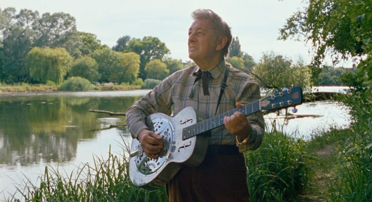 Al Cook steht mit Gitarre vor einem Gewässer in einem Wald.