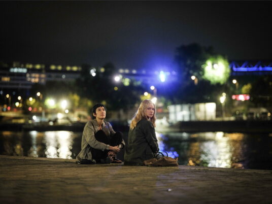 Zwei Personen sitzen am Ufer eines Gewässers bei Nacht vor einer beleuchteten Stadt im Hintergrund