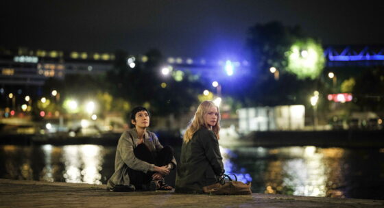 Zwei Personen sitzen am Ufer eines Gewässers bei Nacht vor einer beleuchteten Stadt im Hintergrund